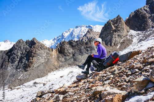 Young female mountaineer in a helmet resting sitting on a backpack and writes the passed route.
