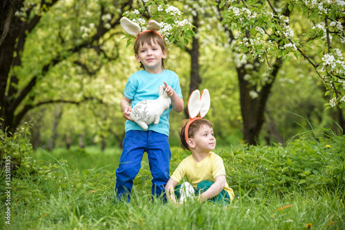 Two little kids boys and friends in Easter bunny ears during traditional egg hunt in spring garden, outdoors. Siblings having fun with finding colorful eggs. Old christian catholoc tradition