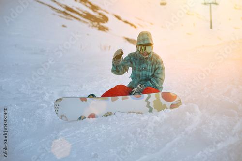 Fallen beginner girl snowboarder wears her google mask, sits alone at traverse of ski slope in sunset rays, smiles and shakes her hand looking at camera, ready to stand up and ride down