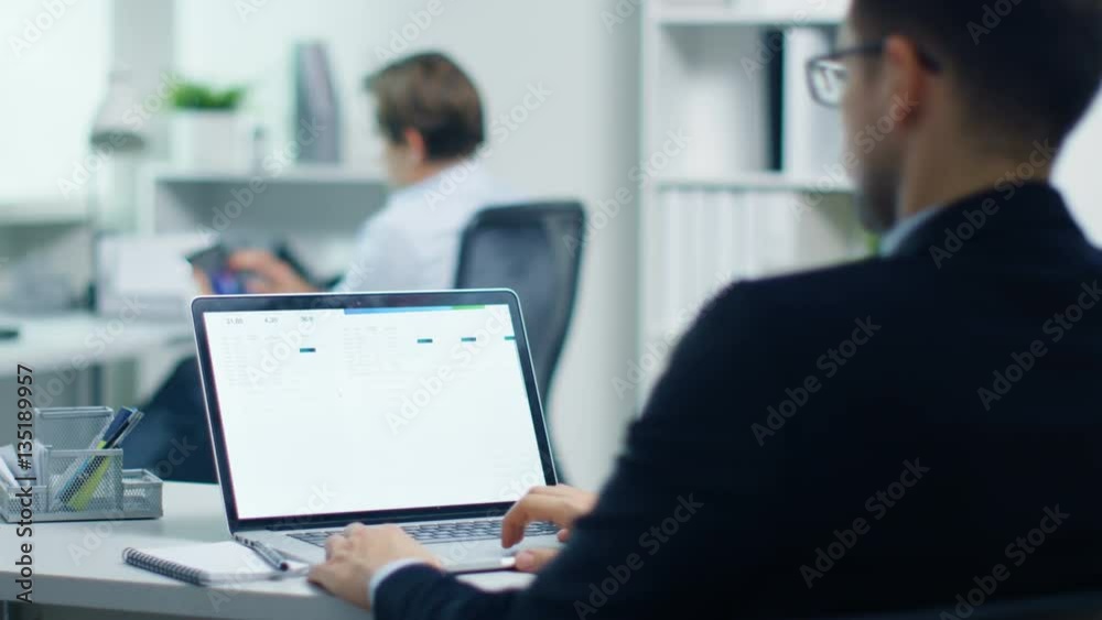 Young Businessman Working with Statistics on His Laptop. His Colleague Sits at His Desk Using Tablet Computer. Shot in a Modern and Light Office. Shot on RED Cinema Camera 4K (UHD). Young Businessman Working with Statistics on His Laptop. His Colleague Sits at His Desk Using Tablet Computer. Shot in a Modern and Light Office. Shot on RED Cinema Camera 4K (UHD).