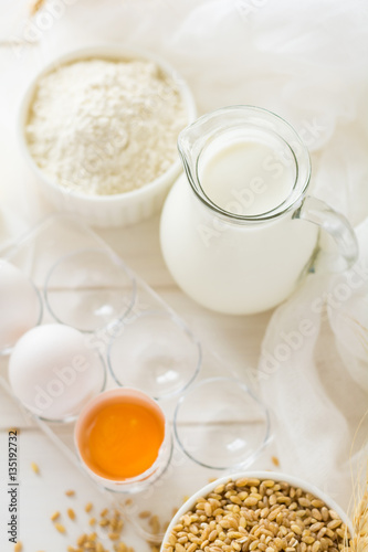 Ingredients for baking: flour, milk, wheat grain, butter and eggs on white wooden background. Selective focus
