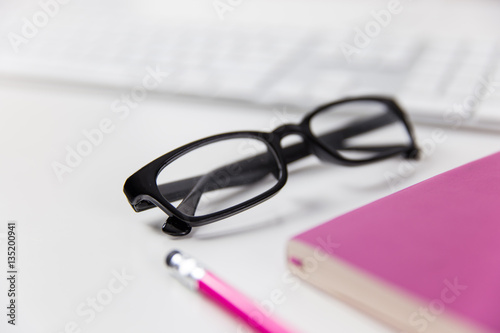 Glasses, pink notebook and computer keyboard on white office desk