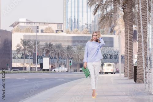 Stylish blond woman in smart casual blouse and white trousers holding a green handbag. Young female walking through empty street in Dubai Downtown  UAE
