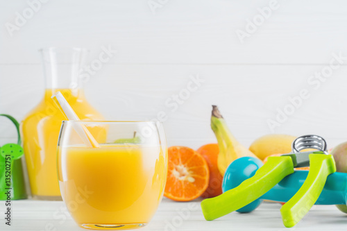 A glass of fresh orange juice and fruits, tangerine, apple, banana, dumbbell, hand gripper on white wooden background. Healthy lifestyle and diet concept. Healthy eating for weight loss. 