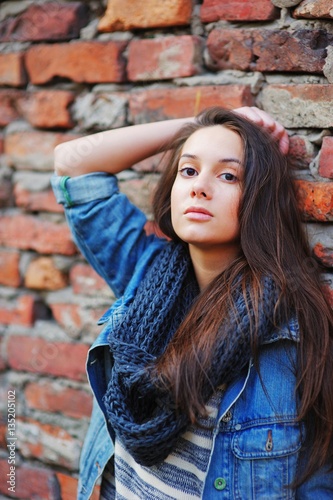 Young pretty girl with long brown hair, dressed in a denim jacket, a striped tee and black scarf looks pensive, standing near a brick wall on the street. © raisondtre