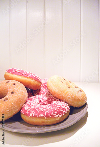 A ring donuts with pastel pink frosting and sprinkles on gray plate. White painted wooden planks on background. Selective focus. Place for your text