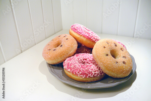 A ring donuts with pastel pink frosting and sprinkles on gray plate. White painted wooden planks on background.