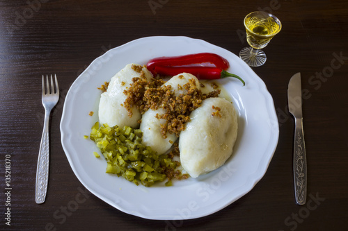 Potato dumplings - a traditional regional dish. Polish and Lithuanian cuisine. With pickles , fried bacon and paprika . White plate, knife, fork and glass of vodka on a dark background..