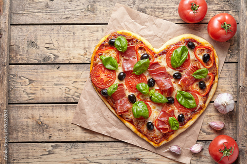 Pizza heart shaped love concept Valentine's Day romantic dinner food. Prosciutto, olives, tomatoes, parsley, basil and mozzarella cheese meal served on vintage wooden table