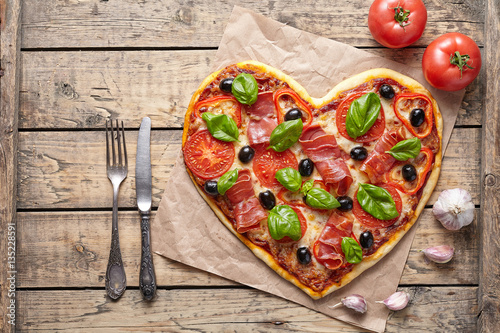 Pizza heart shaped love concept Valentine's Day romantic dinner food with knife and fork. Prosciutto, olives, tomatoes, parsley, basil and mozzarella cheese meal served on vintage wooden table