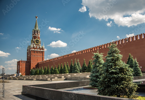 Sunny day at the Red Square with blue sky, white clouds, green fir-trees, the Spasskaya Tower with the Kremlin chimes, the main tower with a through-passage on the eastern wall of the Moscow Kremlin