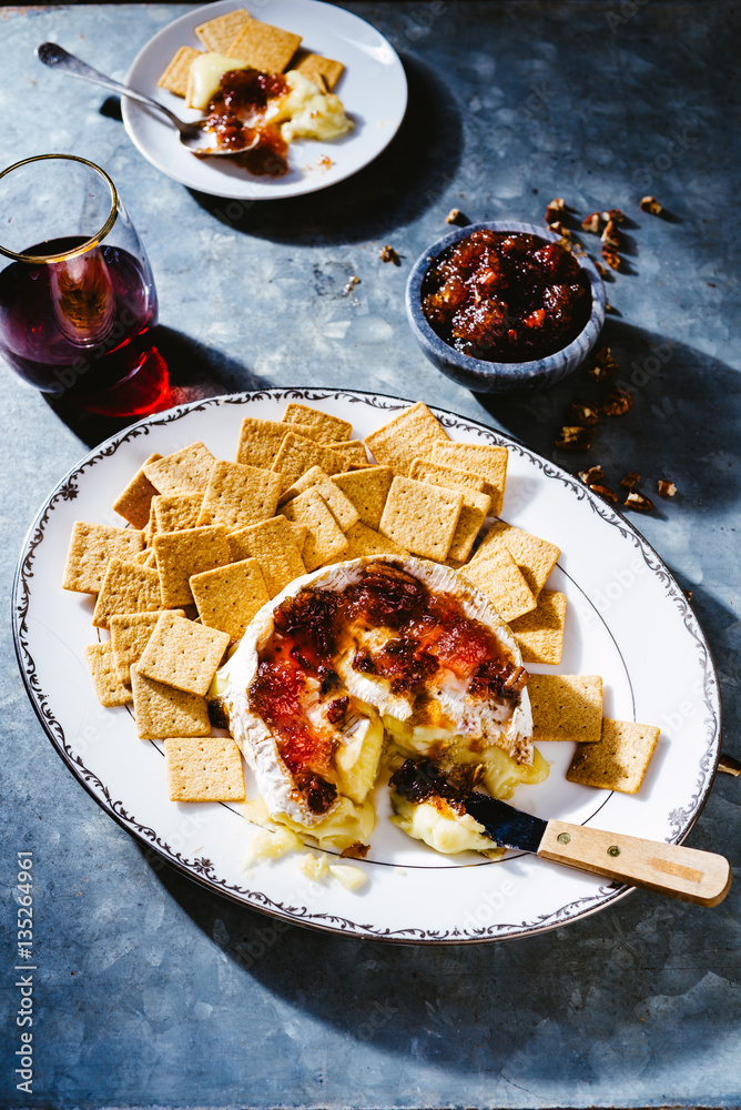 Brie cheese and cranberry pepper jelly party appetizer with crackers and wine on a zinc surface dark and moody lighting. Brie cheese and cranberry pepper jelly party appetizer with crackers and wine on a zinc surface dark and moody lighting.