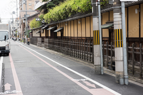 In this image we can see partially see a truck parked by the side of the road. Few green bushes can be seen on the roof of the buildings.
