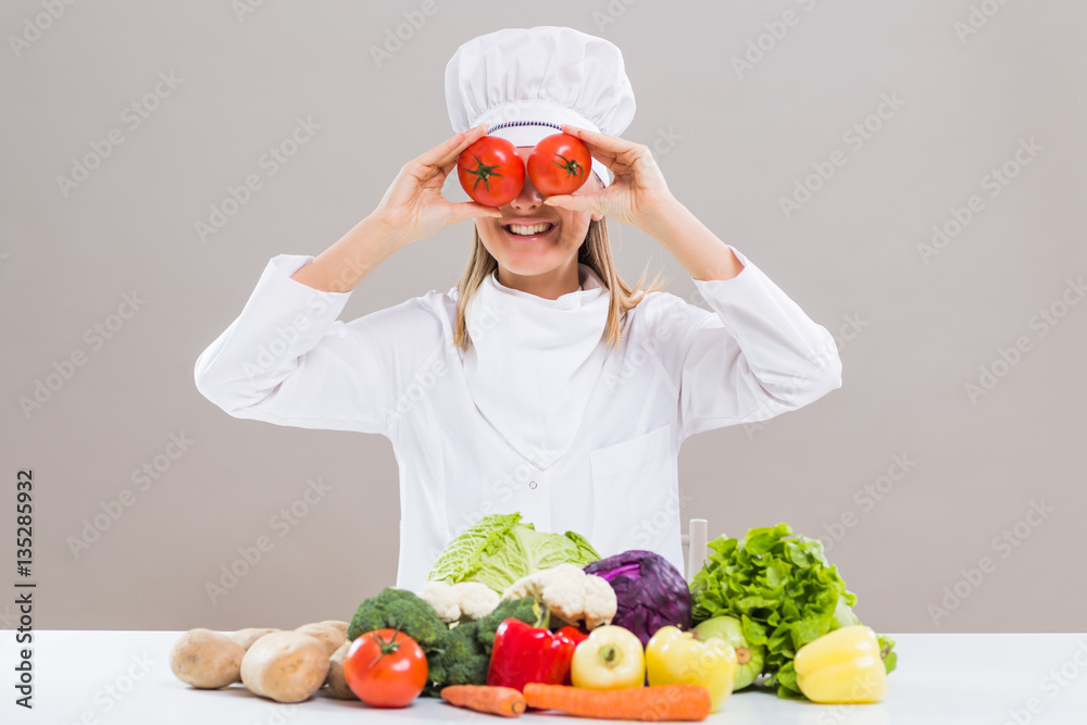 Cheerful female chef is covering her eyes with tomato while sitting at the table with bunch of vegetable.
Cheerful female chef is covering her eyes with tomato while sitting at the table with bunch of vegetable.