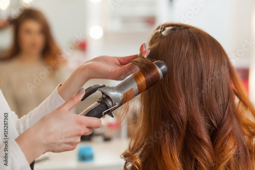 Hairdresser makes hairstyle girl with long red hair in a beauty salon. Create curls with curling irons. Professional hair care.