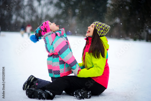 Young woman and the girl in bright ski suits sit having embraced on snow. photo