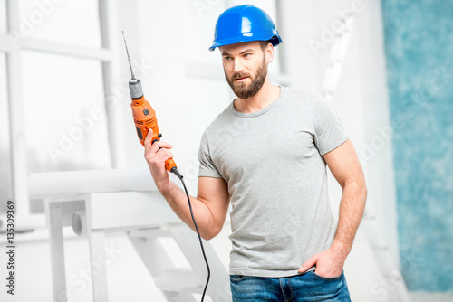 Portrait of a handsome repairman with drill and helmet in the bright interior