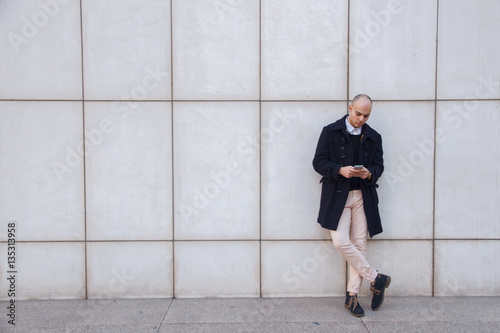 Young handsome bald businessman holding a smart phone, looking down tapping screen leaning on a white wall - technology, business, work concept  © pablobenii