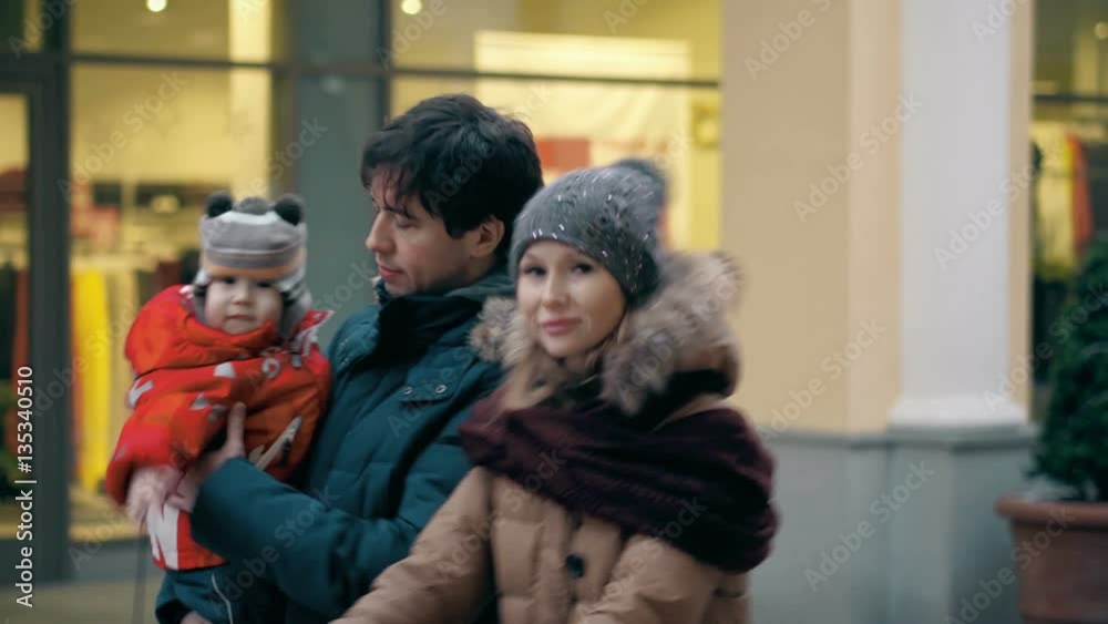 Happy young couple shopping with little baby boy. Parents go on the background of retail shops showcases with discounts signs Happy young couple shopping with little baby boy. Parents go on the background of retail shops showcases with discounts signs