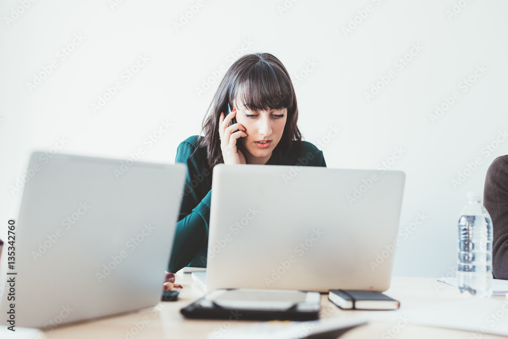 Young beautiful caucasian businesswoman sitting on a desk, using computer talking smart phone - technology, working, business concept Young beautiful caucasian businesswoman sitting on a desk, using computer talking smart phone - technology, working, business concept