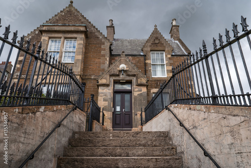 Orkneys, Scotland - June 5, 2012: The facade of Stromness town hall whick stands up a flight of stairs. Brown and gray building under dark cloudy skies. Black fence and gray stone stairs.