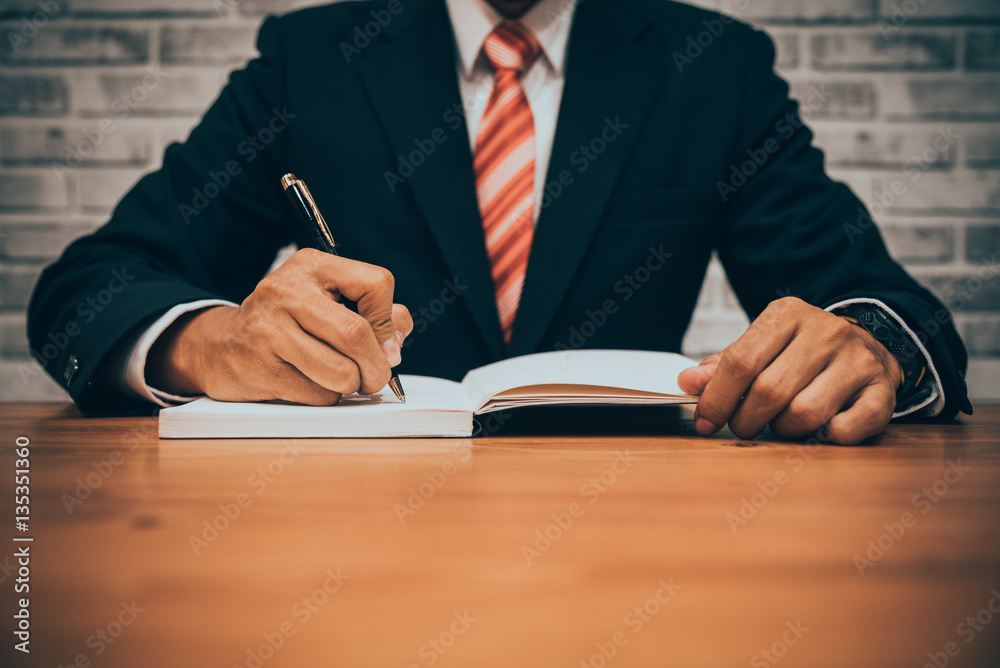 Businessman in suit writing or signing in notebook on wooden tab Businessman in suit writing or signing in notebook on wooden tab