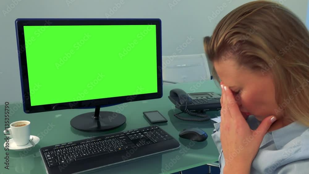 A young, attractive woman sits in front of a computer with a green screen in an office and shakes her head, frustrated