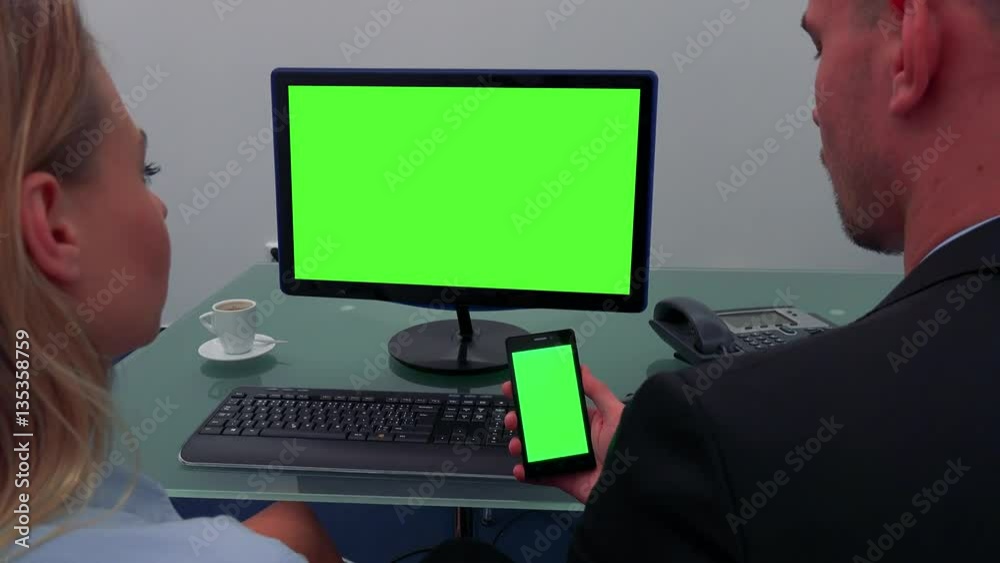 A man and a woman sit at a desk in an office and look in turns at a computer screen and a smartphone (both green screen)