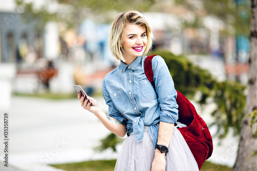A close-up portrait of a standing smiling girl with short blond hair and bright pink lips hoding a smartphone wearing denim shirt, grey tulle skirt, black watch and marsala color backpack