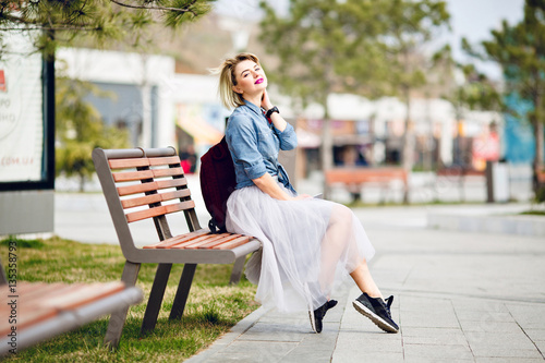 Young cute blond girl with short hair sitting on a wooden bench and  dreaming holding a smartphone and wearing denim blue shirt, grey tulle  skirt, black sneakers and marsala backpack Stock Photo
