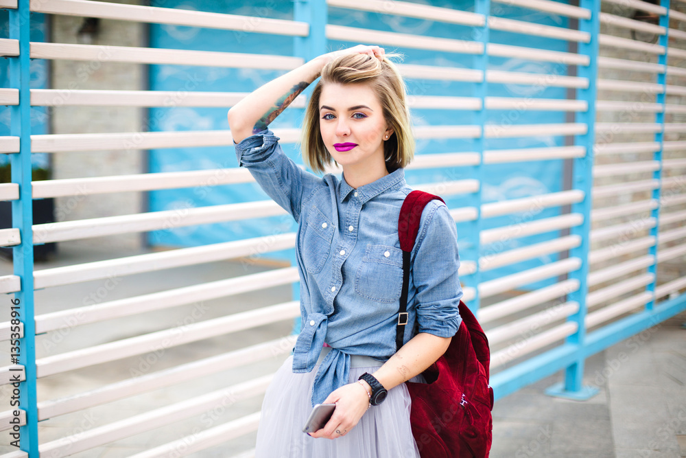 Beatiful tattooed girl with bright pink lips, wearing blue denim shirt, grey tulle skirt and marsala backpack with blue and white stripes on the background