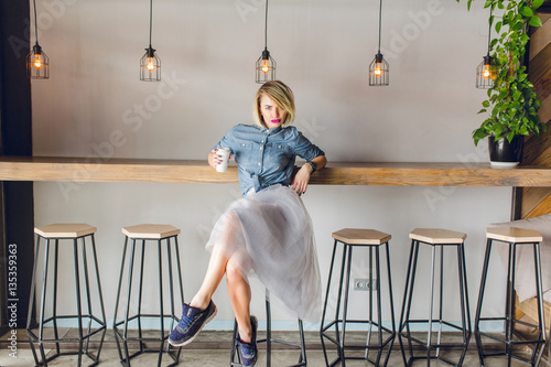 Flirty blond girl with blue eyes and bright pink lips sitting in a coffee shop on a chair drinking coffee. She wears blue denim shirt, grey tulle skirt, black sneakers and watch
