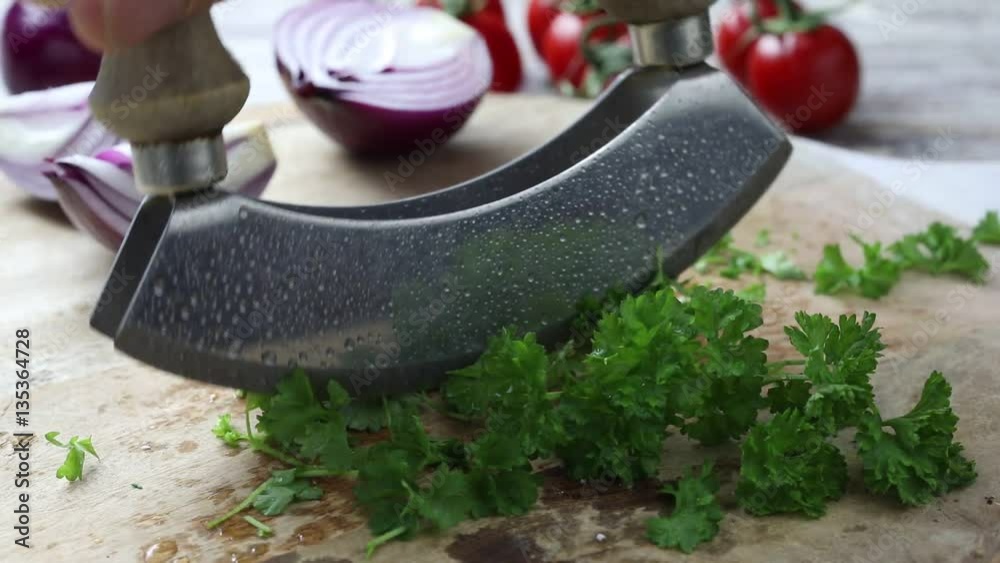 Close up double bladed mezzaluna mincing knife chopping fresh herbs on wooden cutting board with vegetables in soft focus on table