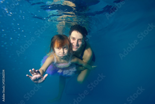 The little girl and the instructor swim underwater, smiling and looking at camera against a blue background. Portrait. Close-up. The view from under the water. Landscape orientation