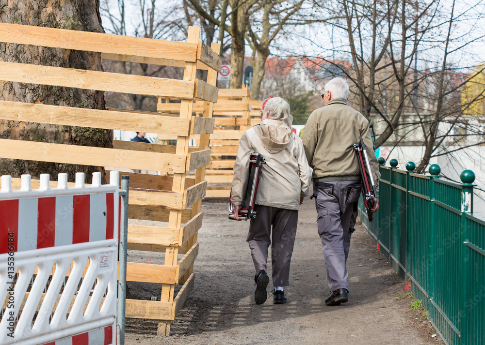 Two seniors - a man and a woman - walking through a park with two folding chairs, taking each other's hand. The couple looking for a nice place to sit and enjoy first warm spring days.