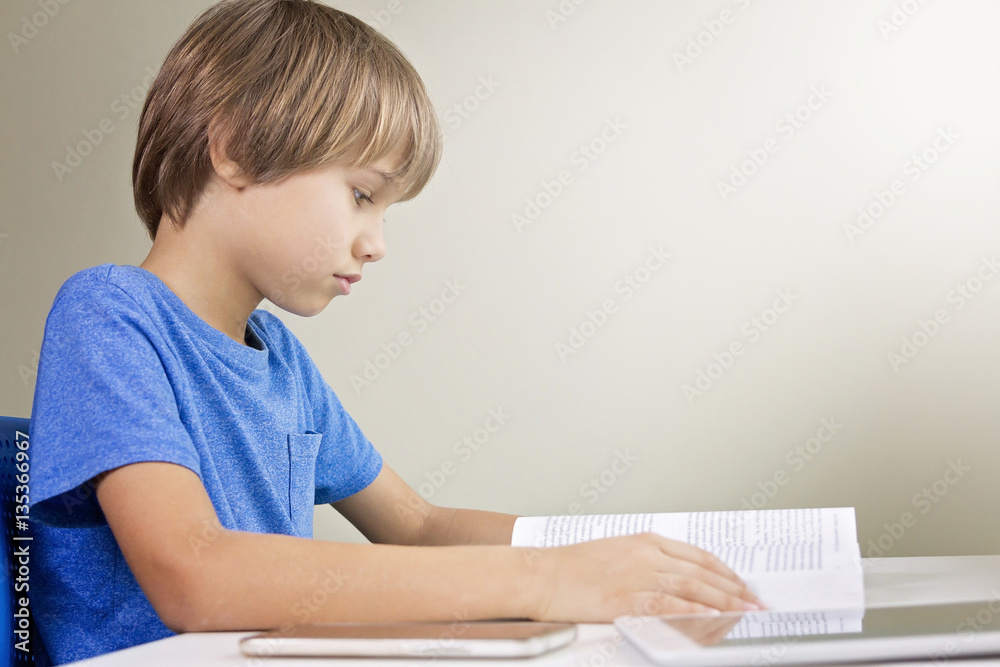 Concentrated child reading a book. Mobile phone and tablet computer are on the table near him. Technology, education, school concept Concentrated child reading a book. Mobile phone and tablet computer are on the table near him. Technology, education, school concept