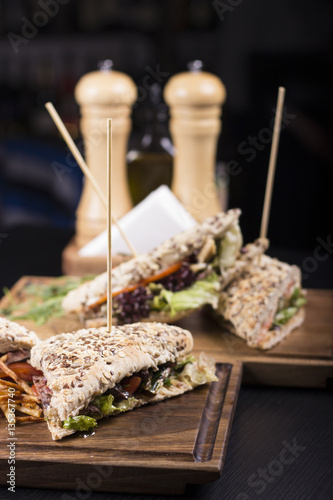 Tasty grilled sandwiches with a wooden stick stabbed trough them on a wooden plate. Meat sandwiches with lettuce  tomatoes and french fries on the side. Napkins and olive oil on the background.