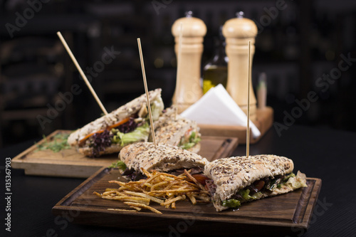 Tasty grilled sandwiches with a wooden stick stabbed trough them on a wooden plate. Meat sandwiches with lettuce, tomatoes and french fries on the side. Napkins and olive oil on the background.