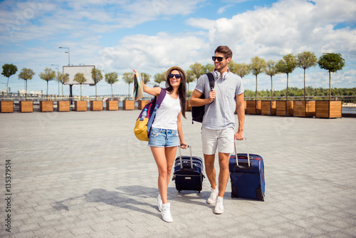 Two smiling young lovers with valise walking from airport in to