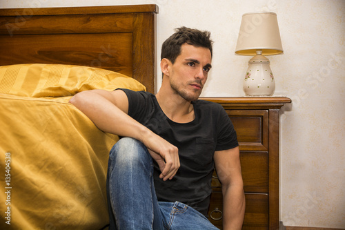 Handsome young man sitting next to his bed with serious expression, staring in the void with a blank look