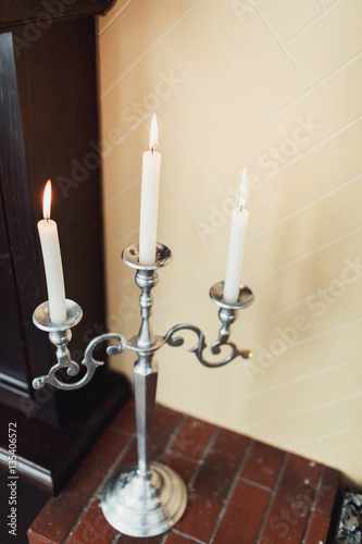 Silver candleholder with white candle stands by wooden fireplace