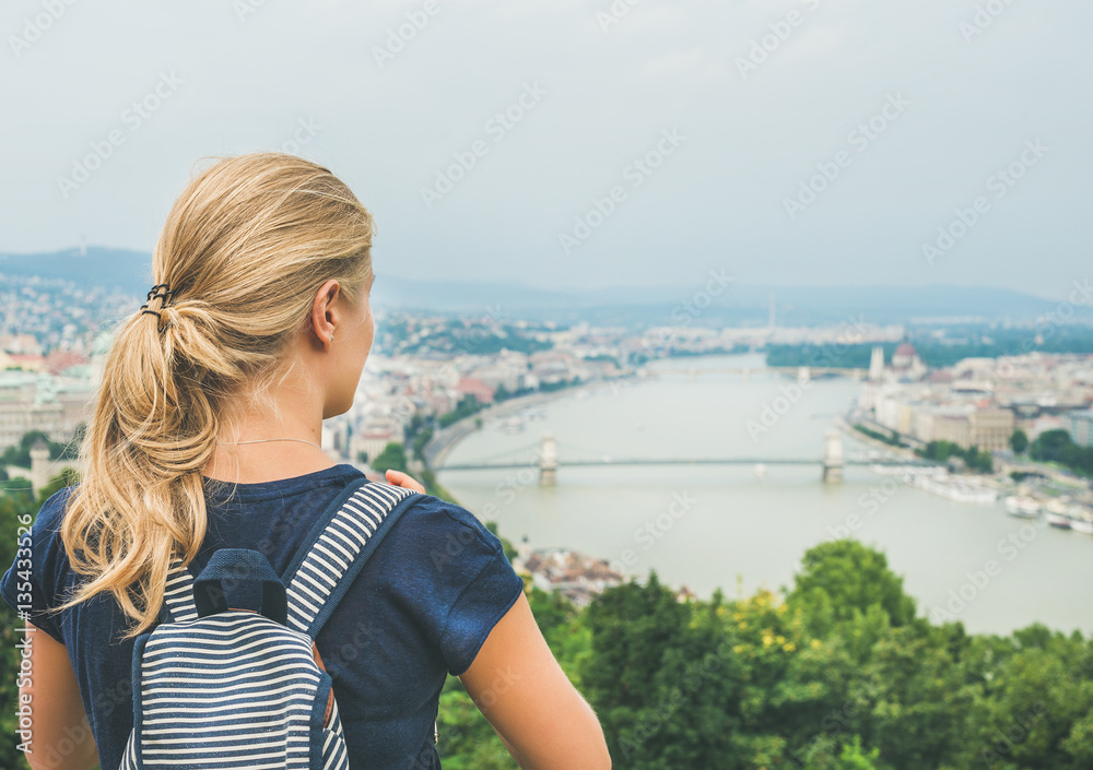 Young blond woman traveler with backpack looking at Danube and Budapest river from hill, rear view, Hungary