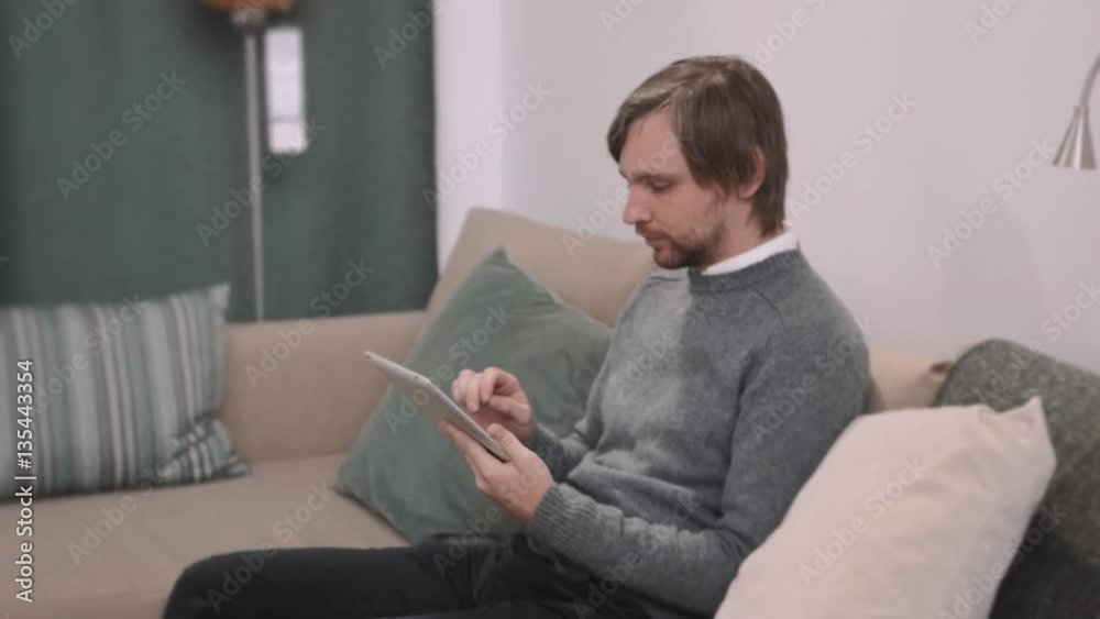 Handsome young man using his digital tablet with smile while lying down on the couch at home. Spending great time at home. Handsome young man using his digital tablet with smile while lying down on the couch at home. Spending great time at home.