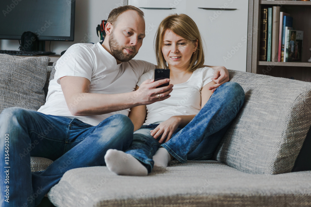 Natural, lifestyle image of attractive couple sitting on couch together looking at smartphone in the living room.