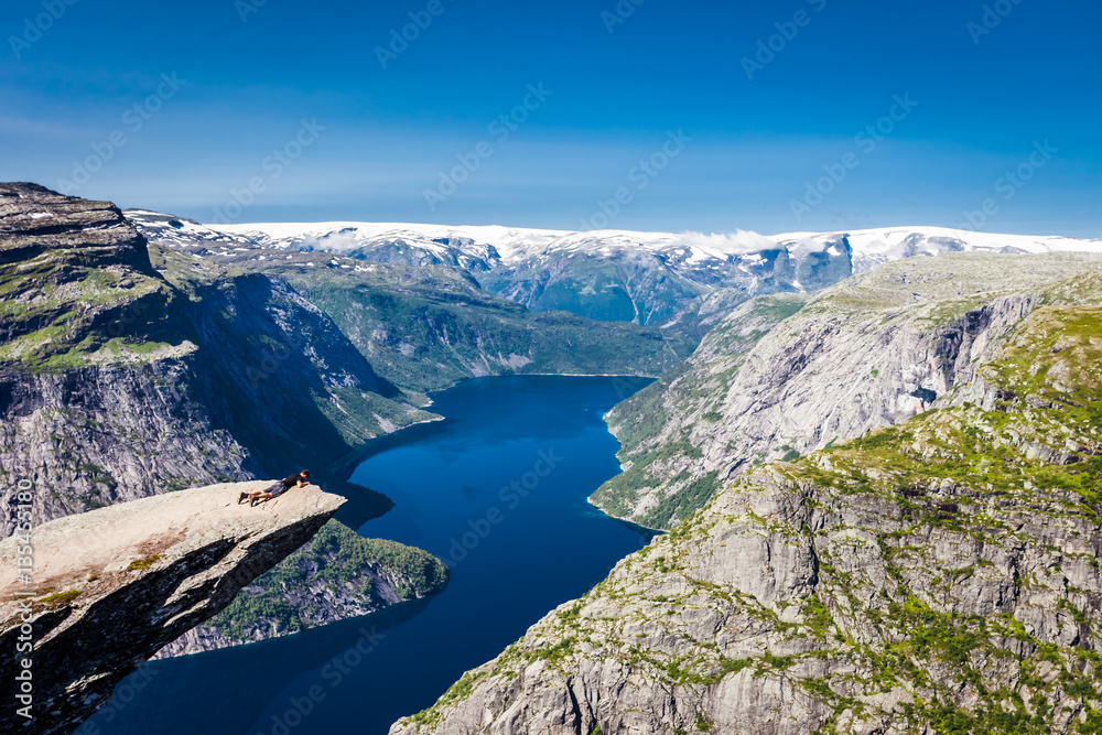 Panoramic view of lying man on Trolltunga cliff, Norway, Scandinavia. Sunny day with amazing blue sky. Majestic mountains in the background. Clear blue water. 
