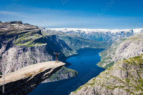 Panoramic view of standing man on Trolltunga cliff, Norway, Scandinavia. Sunny day with amazing blue sky. Majestic mountains in the background. Clear blue water. 
