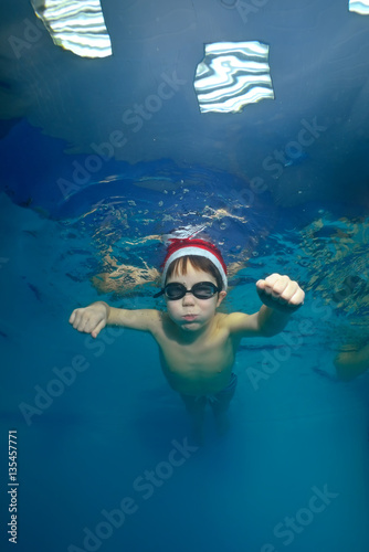 A little boy goes in for sports in the pool  swims underwater in a beanie and glasses and looking at the camera. Bottom view from under the water. Portrait. Vertical orientation