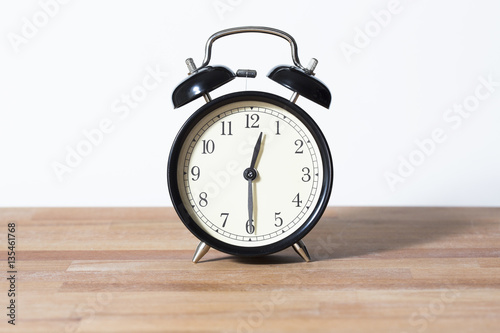 It is half past twelve o'clock. The time is 12:30. Retro clock isolated on a wooden table. White background.