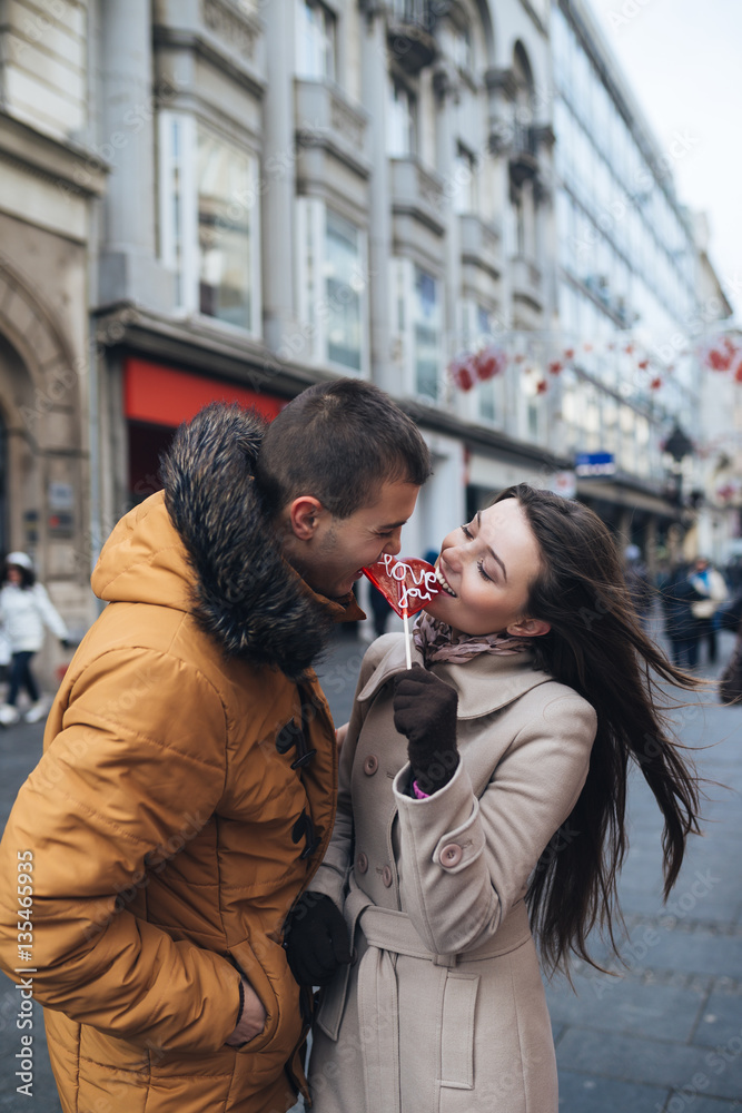 Fototapeta premium Beautiful young couple in love enjoying together at city street, kissing and holding together a lollipop in heart shape. 