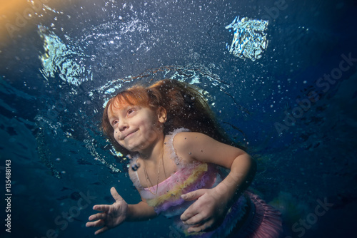Happy little girl floats underwater in a beautiful gown the rays of light and smiling. Portrait. Shooting under the water surface. The landscape view